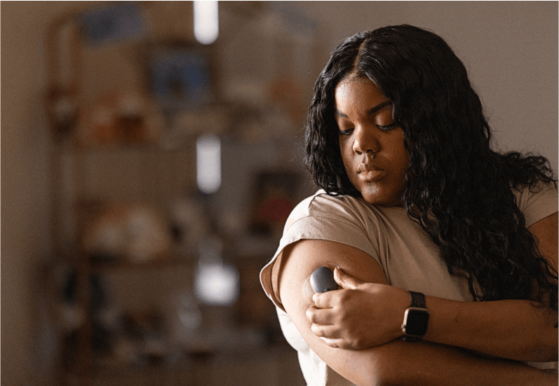Person applying a medical device to upper arm, foreground focus, in a softly lit room with shelves in background.